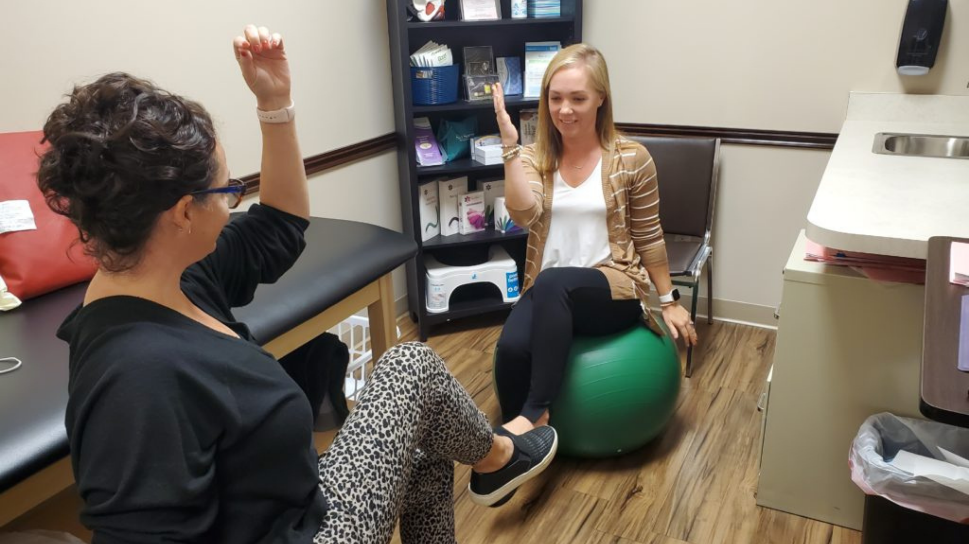 Woman sitting on a green bouncy ball with her right arm bent at 90 degrees up, repeating after her physical therapist.